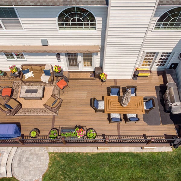 Top-Down Aerial View Of A Large Brown Composite Deck Featuring A Lounge Area With Fire Pit Table Dining Table With Umbrella Grill Station And Perimeter Railings With Potted Flowers