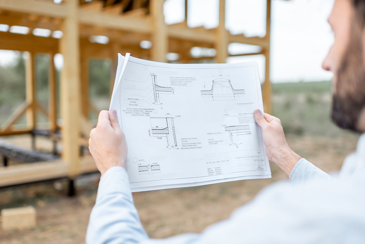 Professional Contractor Holding Technical Blueprints While Standing In Front Of A Wooden Frame Structure, Demonstrating How To Build A Sunroom On A Deck.