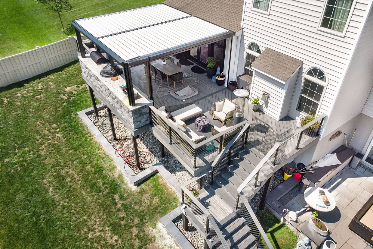 High-Angle View Of A Luxury Multi-Level Deck Featuring A White Louvered Pergola Over The Dining Area, Illustrating Covered Deck Design For Year-Round Outdoor Living.