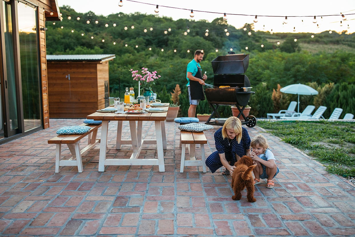 A Family Enjoying A Summer Evening On A Red Brick Patio With Overhead String Lights And A Dining Table, Illustrating Family-Friendly Patio Ideas And Functional Outdoor Living Space Usage.