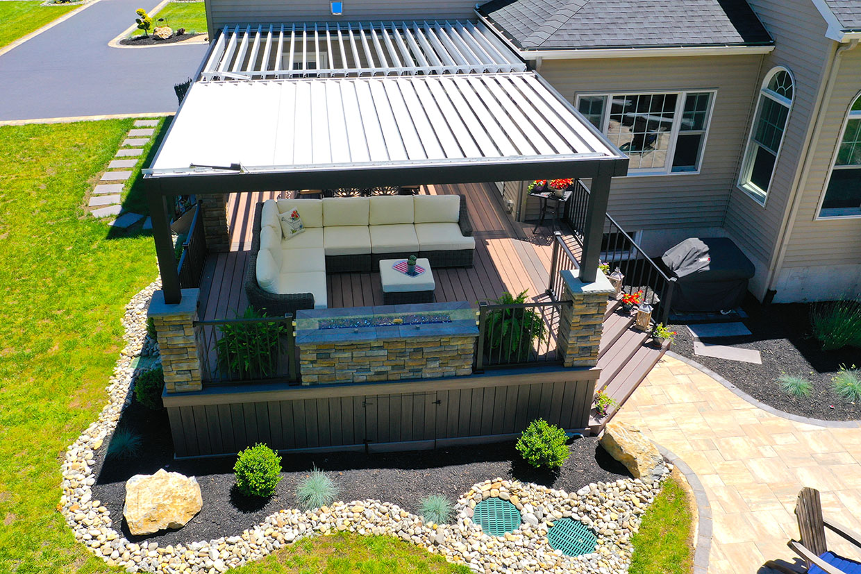 High-Angle View Of An Elevated Custom Composite Deck With A Black Louvered Pergola Stacked Stone Columns Cream Sectional Sofa Built-In Stone Fire Feature And Vertical Composite Skirting