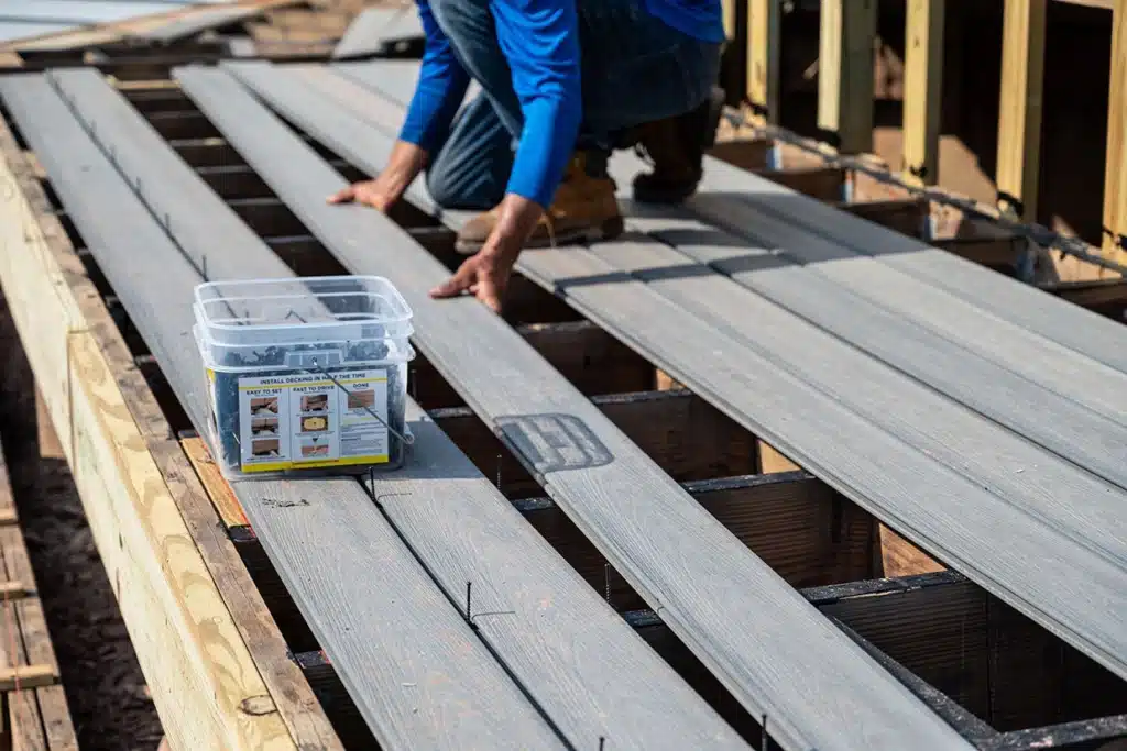 A worker installing decking boards on a new deck framework using a container of deck fasteners. DIY vs Professional Deck Building.
