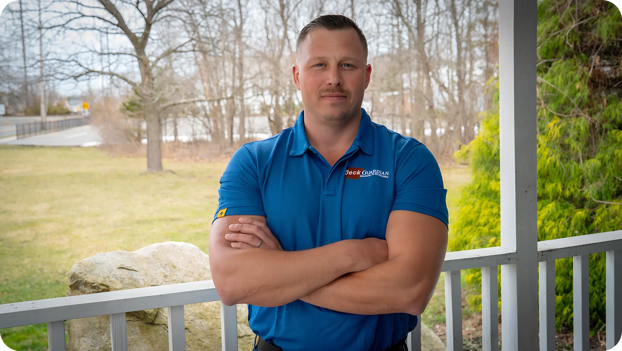 Confident Man In A Blue Deck Guardian Polo Shirt Standing On A Porch With Arms Crossed.