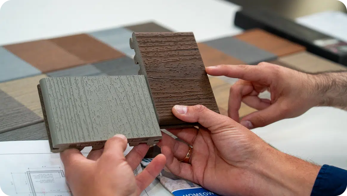 Hands Holding Grey And Brown Composite Decking Boards Over Architectural Blueprints, Comparing Wood-Grain Textures.