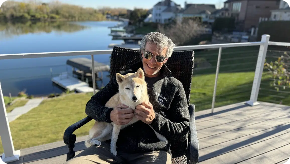 A Smiling Man In Sunglasses Sits In A Patio Chair On A Modern Deck, Holding A Small Cream-Colored Dog On His Lap.