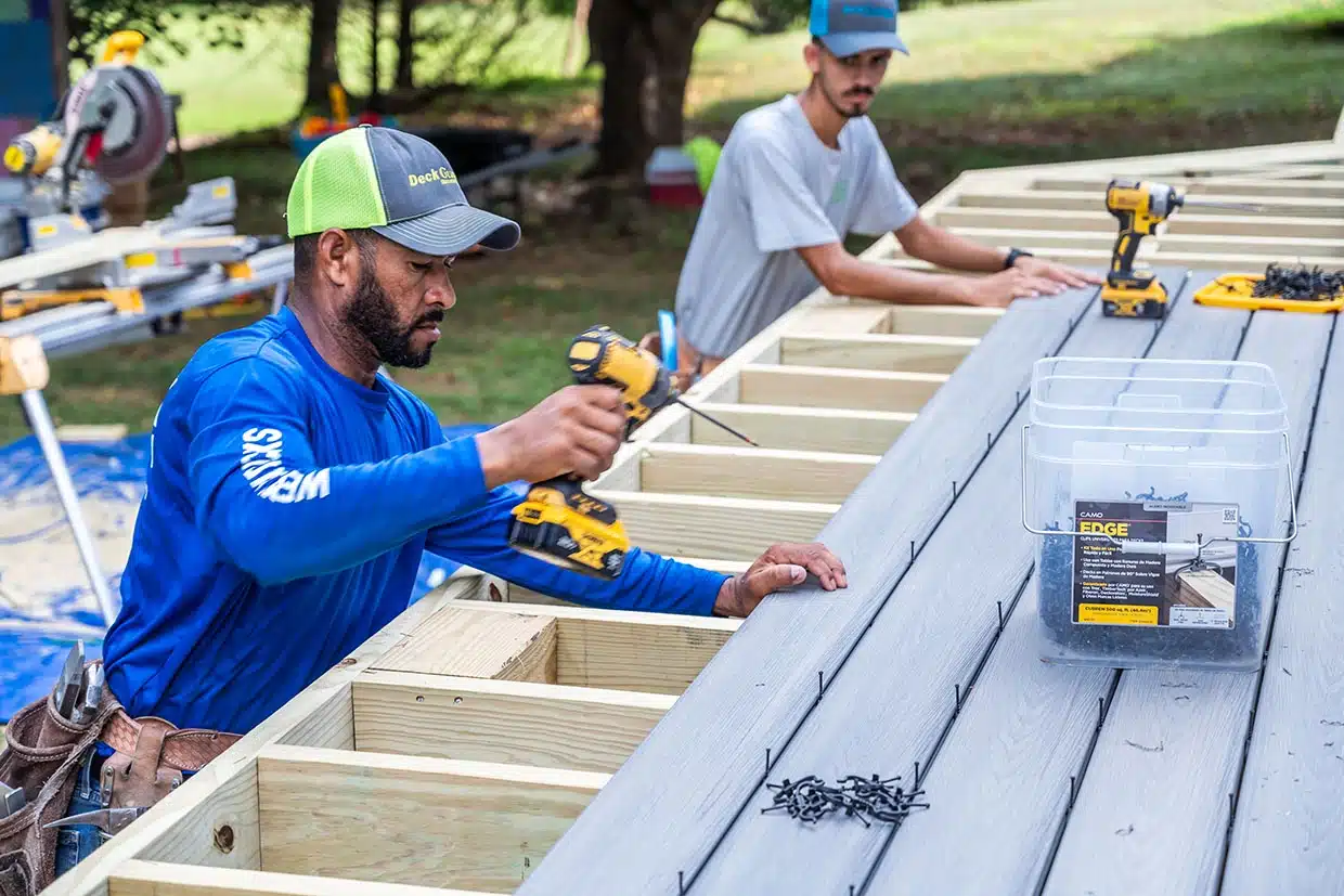 Two Contractors Installing Grey Composite Deck Boards Onto A Wooden Frame Using Power Drills Outdoors.